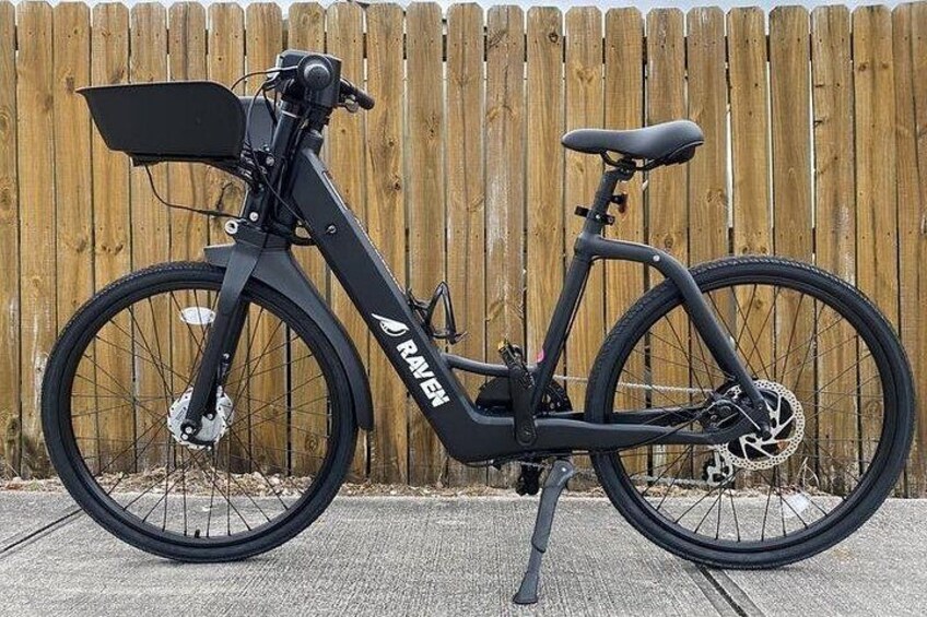 Una bicicleta eléctrica negra con canasta, marca 'RAVEN', estacionada frente a una valla de madera. 4