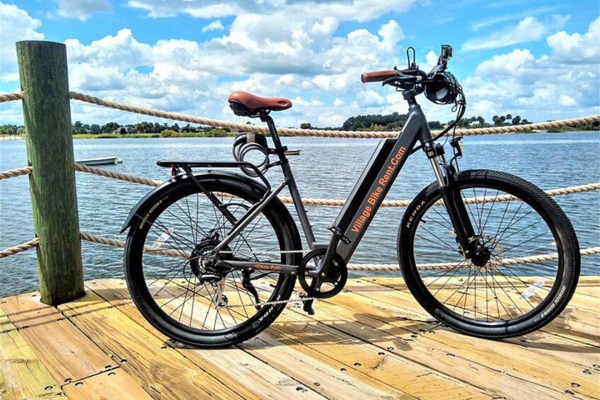 Una bicicleta eléctrica negra con asiento rojo y neumáticos negros, estacionada sobre un muelle de madera junto a un cuerpo de agua. 2