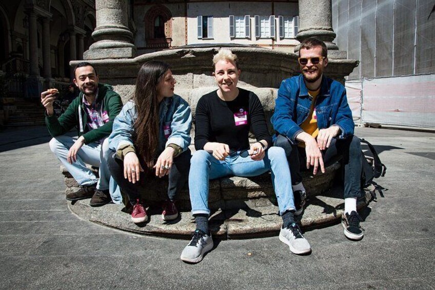 Four people sitting on a stone bench in an outdoor setting with a fountain and classical architecture in the background. 5