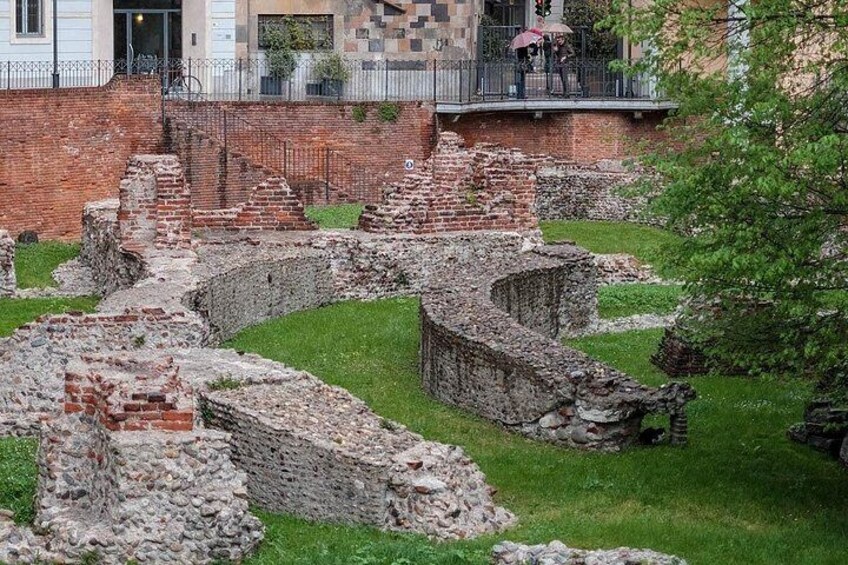 Ancient ruins with brick walls and stone structures, a grassy area, and a building in the background. 2