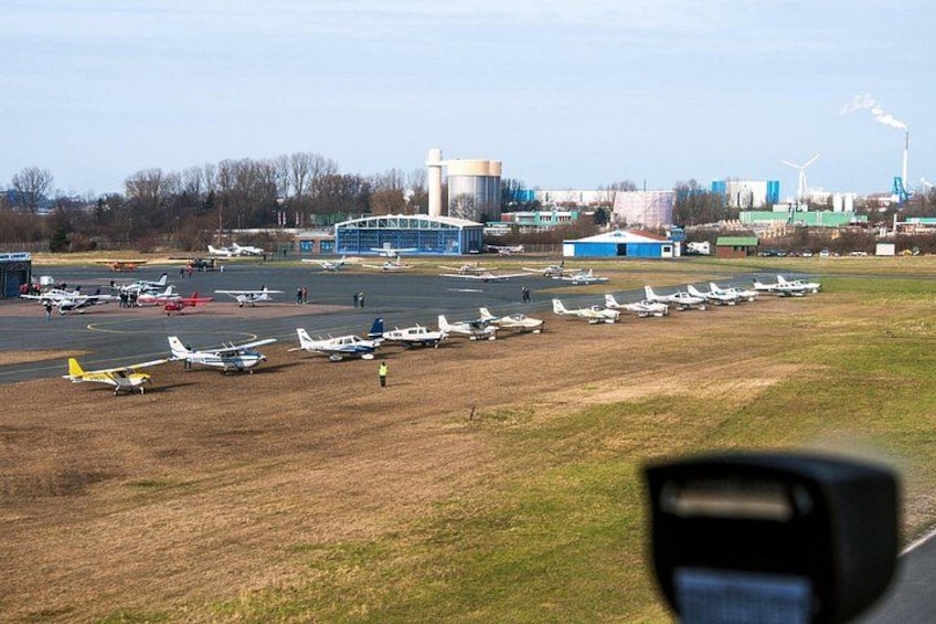An airport with multiple small aircraft parked on the tarmac, a large building in the background, and wind turbines in the distance. 4