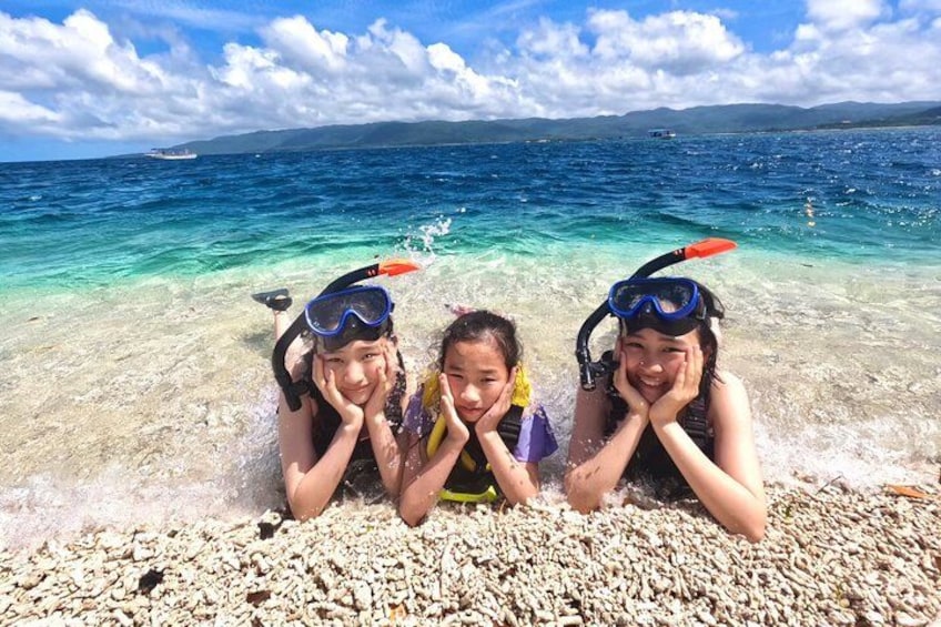 Three people snorkeling in clear blue water with a rocky beach and distant mountains. 2