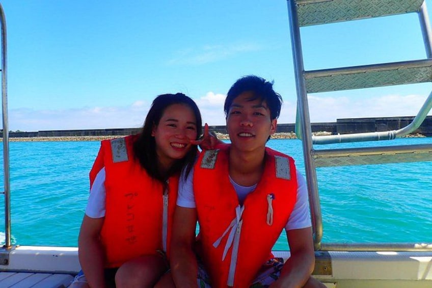 Two people wearing life jackets on a boat with a clear blue sea and a rocky shoreline in the background. 5