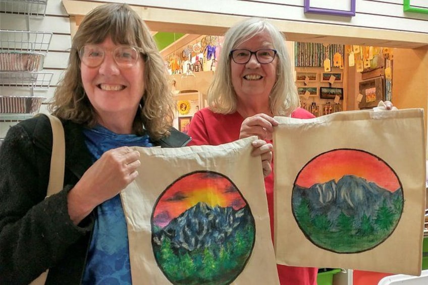 Two individuals holding up painted canvases with mountain landscapes. 2