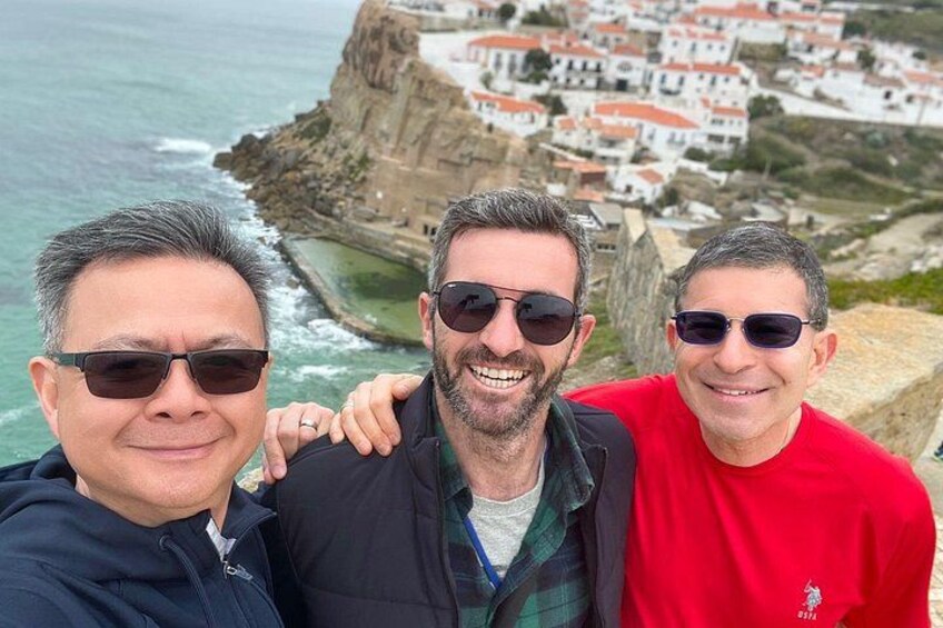 Tres personas en un risco con vista a un pueblo costero con edificios blancos y terreno rocoso. 2