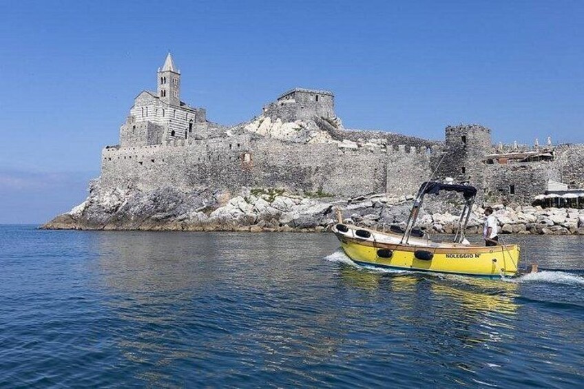 A historic stone fortress with a bell tower, a yellow boat with 'Voleggio N' on the side, and calm blue waters. 2