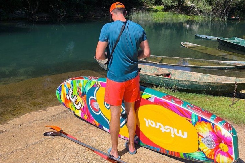 A person standing by a river with a colorful paddleboard, boats, and a calm water surface. 4