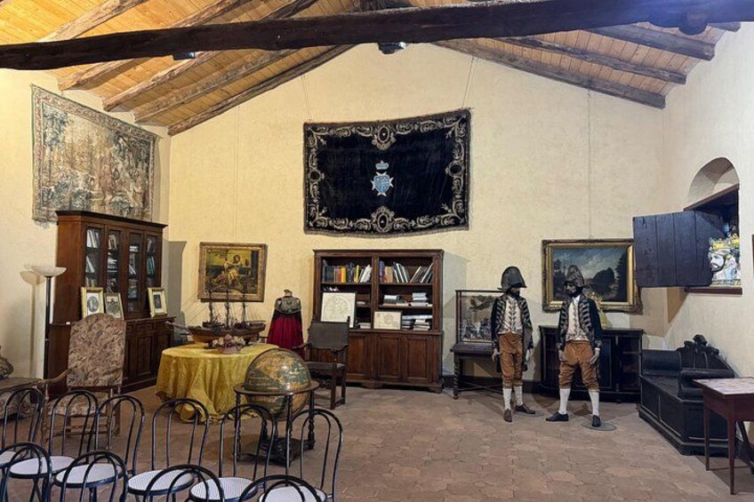 Una habitación con techo alto, vigas de madera, un tapiz grande, un mueble de madera, una mesa con mantel amarillo y dos maniquíes vestidos con trajes de época. 5