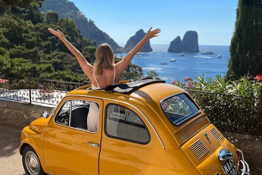A vintage yellow car parked by a coastal road with a woman standing on top, arms raised, and a scenic view of the sea and mountains in the background. 2