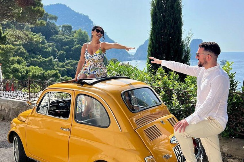 A yellow vintage car with a woman standing on the roof and a man pointing towards the sea. 4
