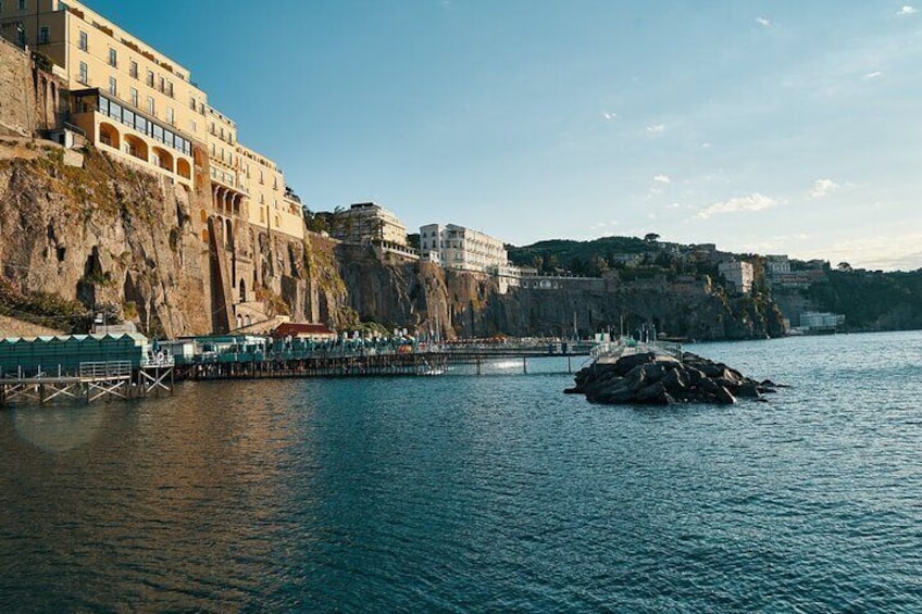 A coastal town with buildings on a cliff, a pier extending into the water, and a rocky outcrop in the center. 4
