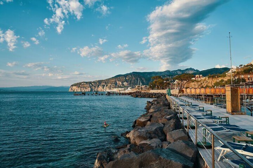 A coastal town with a rocky breakwater, clear blue sea, and a distant mountain range. 5