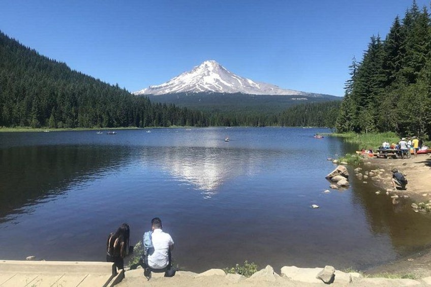 Un lago con una montaña al fondo, una persona sentada en la orilla, y un grupo de personas a la derecha. 2
