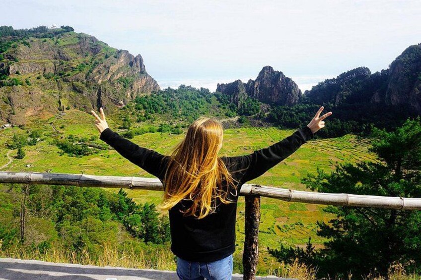 A person with arms outstretched, overlooking a mountainous landscape with greenery and rocky outcrops. 5