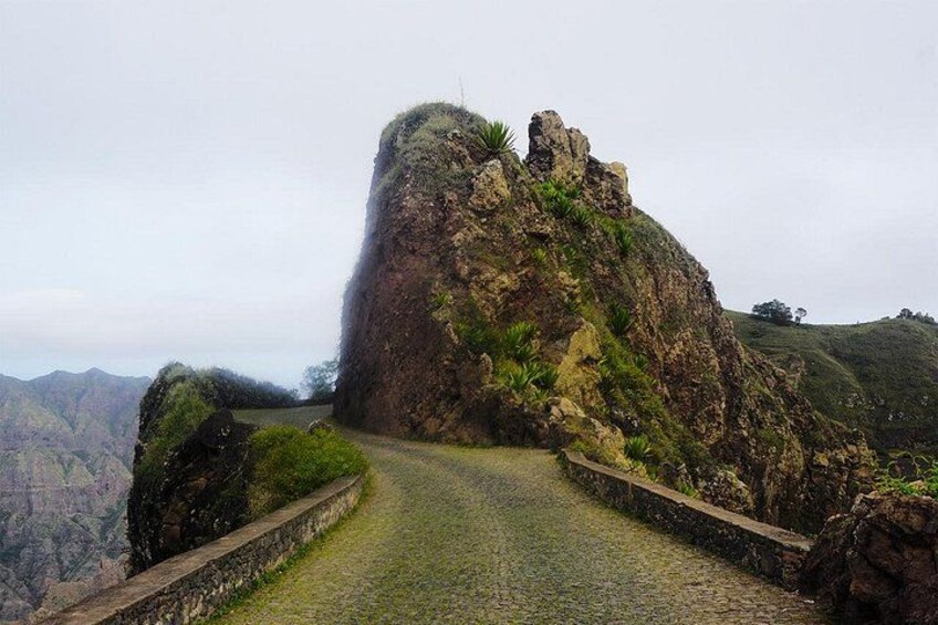 A rugged mountain landscape with a narrow, winding road leading to a large rock formation. 3