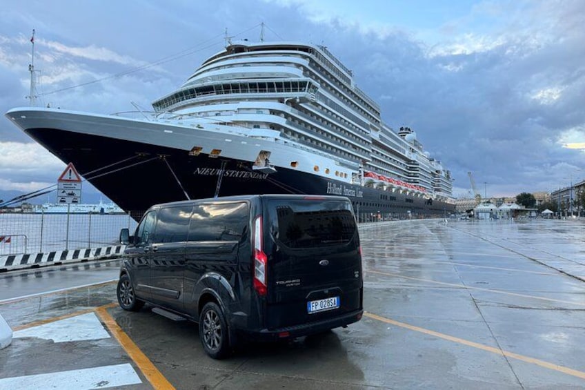 A black van parked on a wet dock with a large cruise ship in the background. 1