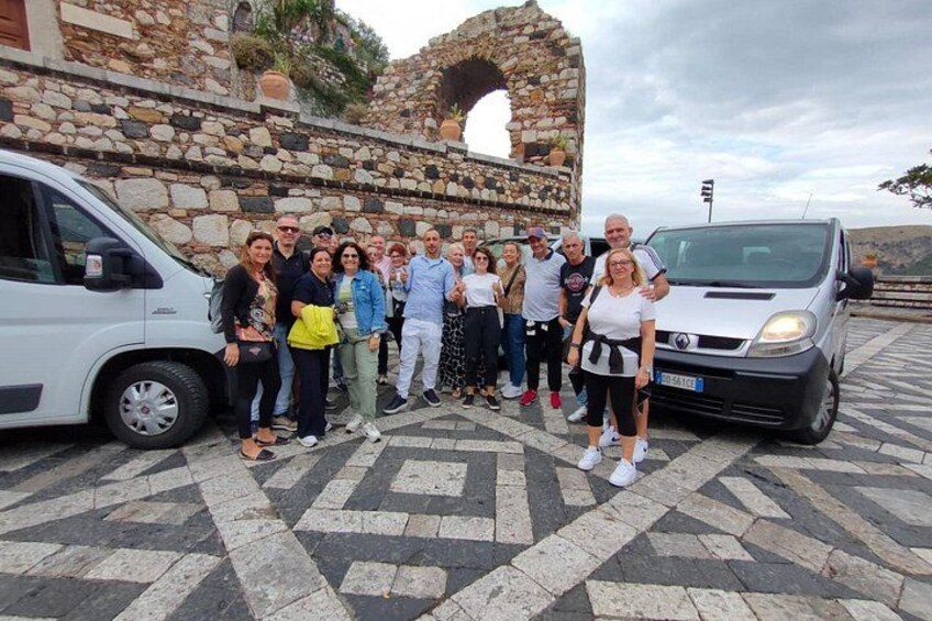 A group of people standing in front of a stone building with a large archway, a white van, and a silver car parked nearby. 1