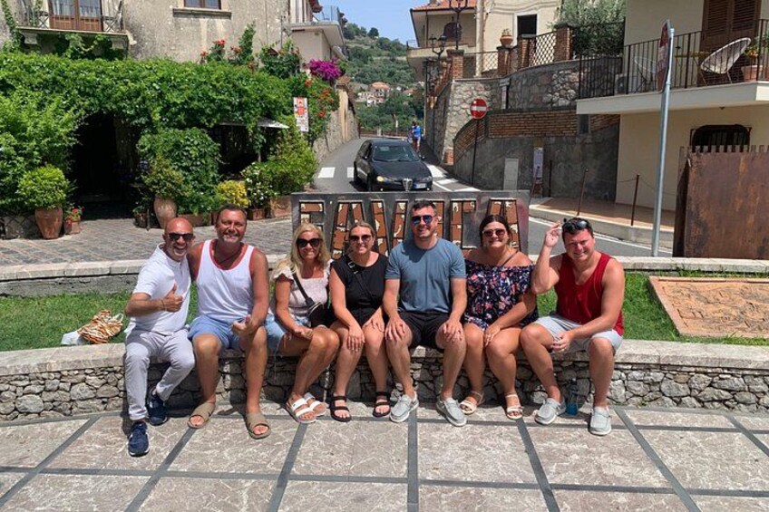 A group of people sitting on a stone bench in a sunny outdoor setting with a stone wall and greenery in the background. 3