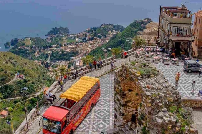 A red bus on a winding road with a yellow canopy, a stone wall, and a view of a coastal town. 2