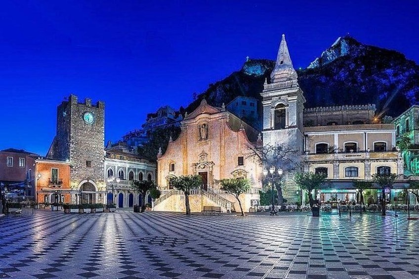 A historic square with a clock tower, a church, and a mountain backdrop. 4