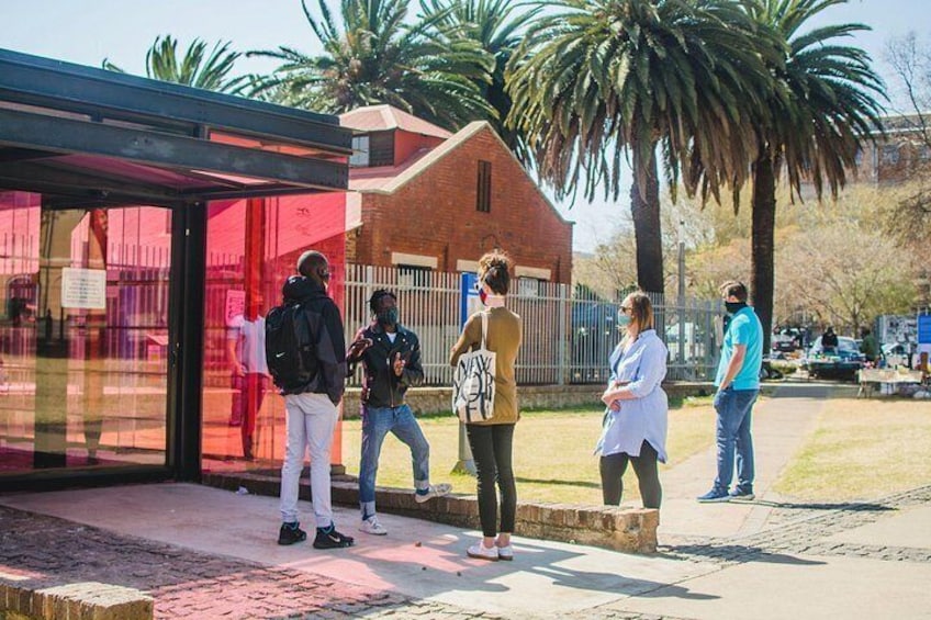 A group of people standing near a bus stop shelter with palm trees and a brick building in the background. 4