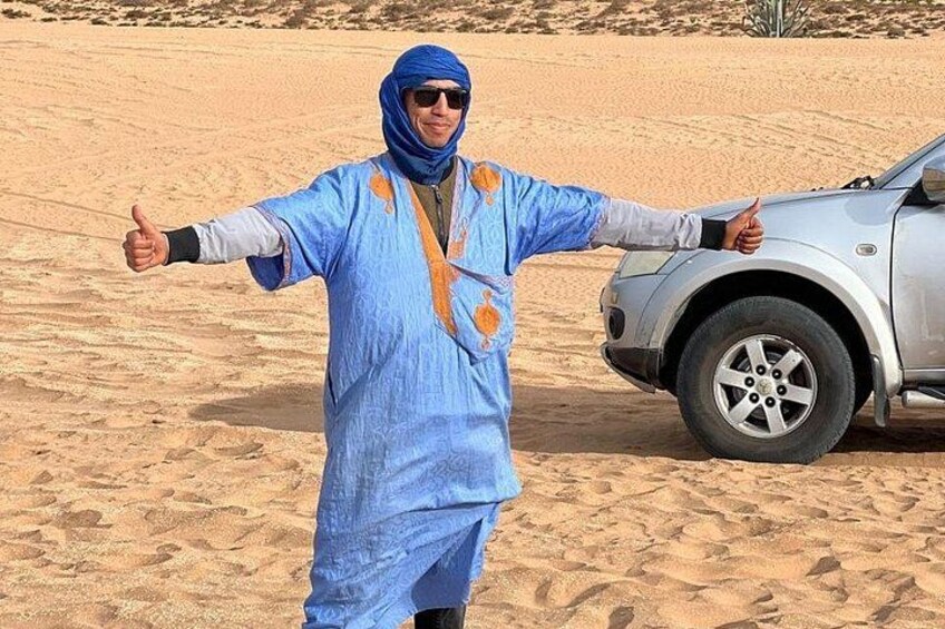 A person in traditional attire giving a thumbs-up gesture in a desert landscape with a car nearby. 1