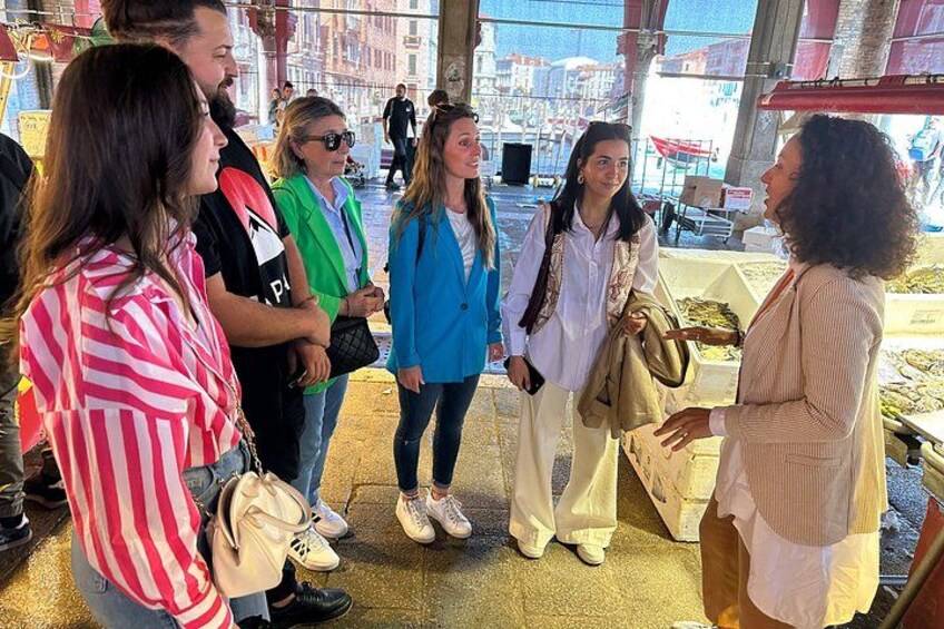 Eine Gruppe von Menschen steht draußen, möglicherweise auf einem Markt oder bei einer öffentlichen Veranstaltung, mit einem Gebäude und einer Straße im Hintergrund. 5