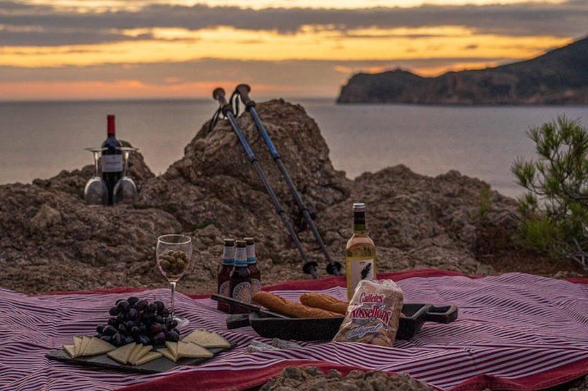 A picnic setup with food, drinks, and a wine bottle on a blanket by a rocky coastline at sunset. 4