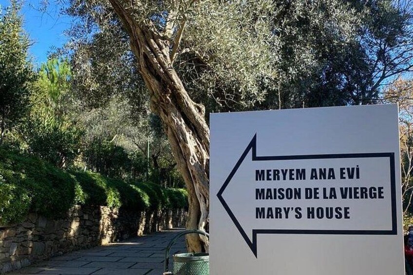 En vejskiltevise for 'Meryem Ana Evı Mansion de la Vierge Marys Hus', med en pil der peger mod venstre. 3