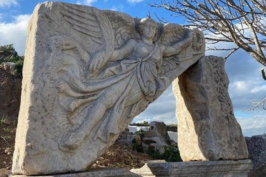 En stor stenskulptur med en figur med vinger, foran en klar himmel og et fjerntliggende landskab. 4
