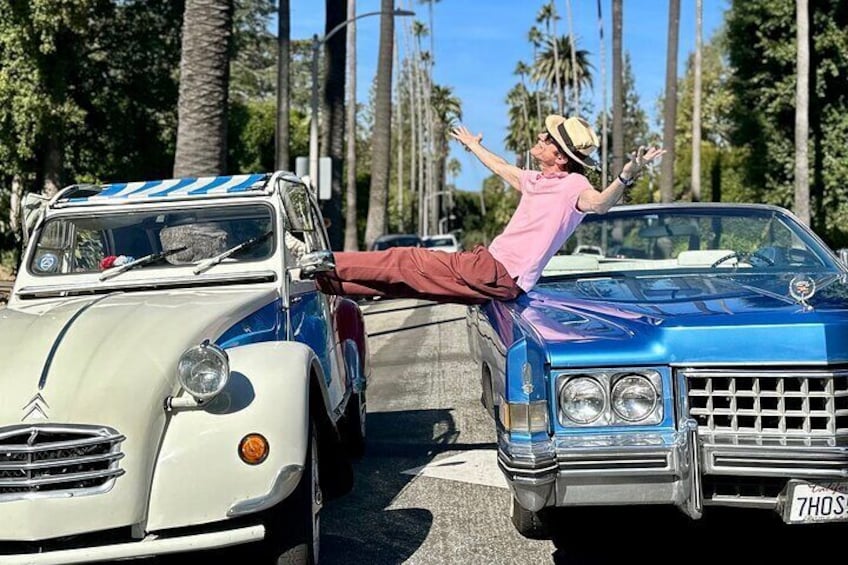 Una persona con camisa rosa y pantalones rojos asoma la cabeza por la ventana de un coche azul, con otro coche blanco estacionado a un lado. 2
