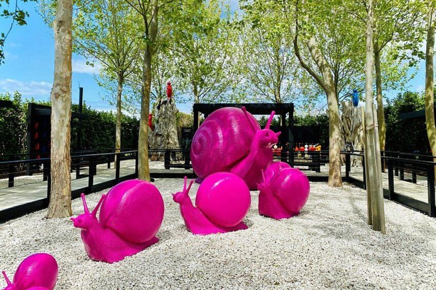 Stora, rosa skulpturer föreställande sniglar i en park med träd och en lekplats i bakgrunden. 2