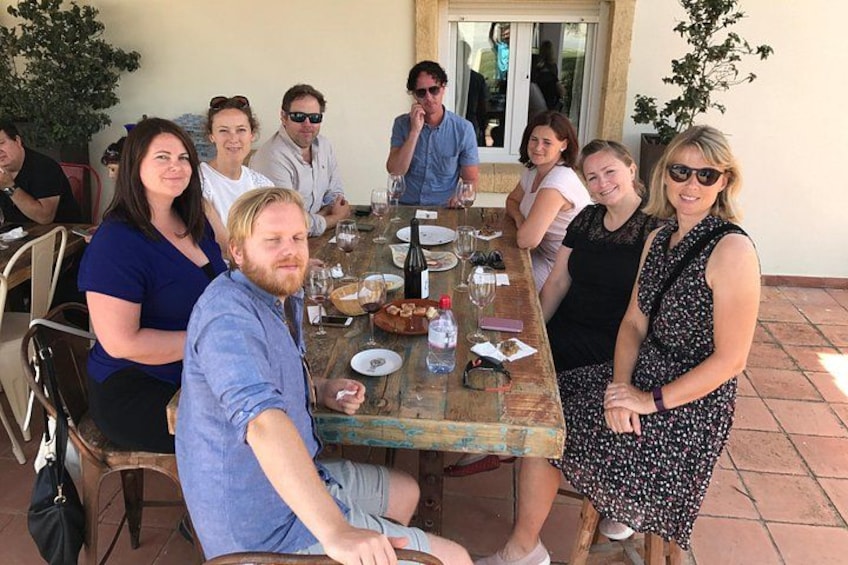 Un groupe de personnes est assis autour d’une table en bois à l’extérieur, et des plats et des boissons sont disposés sur la table. 1