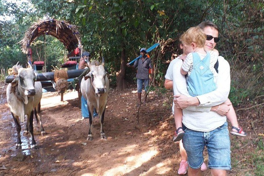 A person carrying a child, walking with cattle, and a cart in a rural setting. 1
