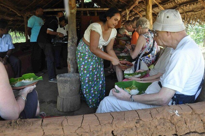 People seated outdoors, some holding plates with food, under a thatched roof structure. 2