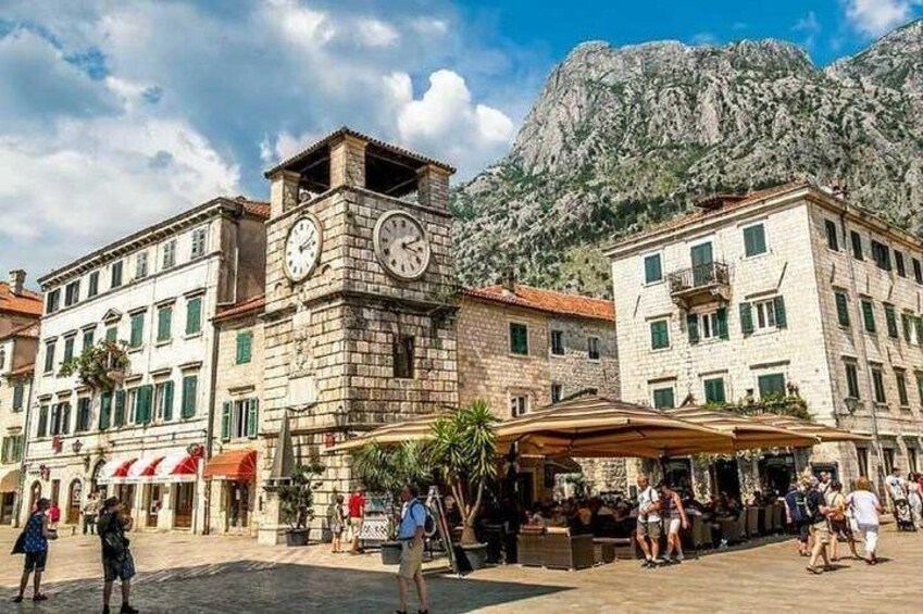 A historic square with a clock tower, stone buildings, and a mountainous backdrop. 1