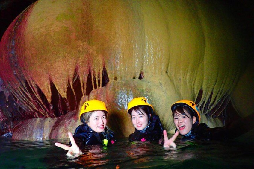 Three people in a cave with yellow helmets, water, and stalactites. 3