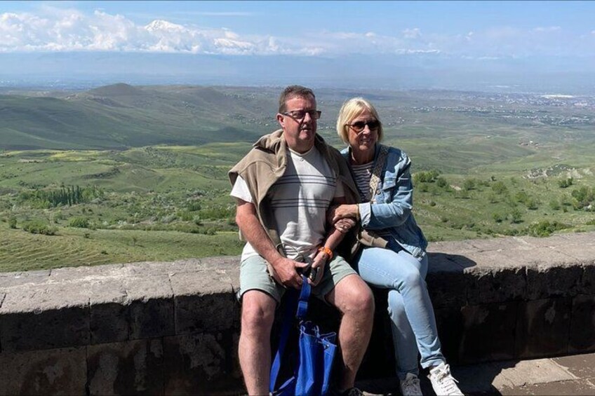 Two people sitting on a stone wall overlooking a green landscape with hills and a distant town. 2