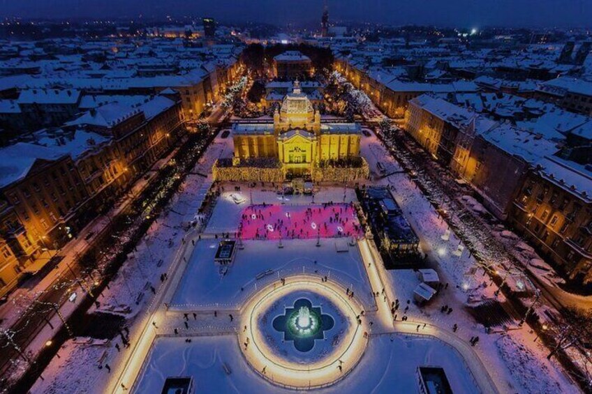 A snow-covered plaza with a central fountain and surrounding buildings, illuminated by artificial lighting. 3