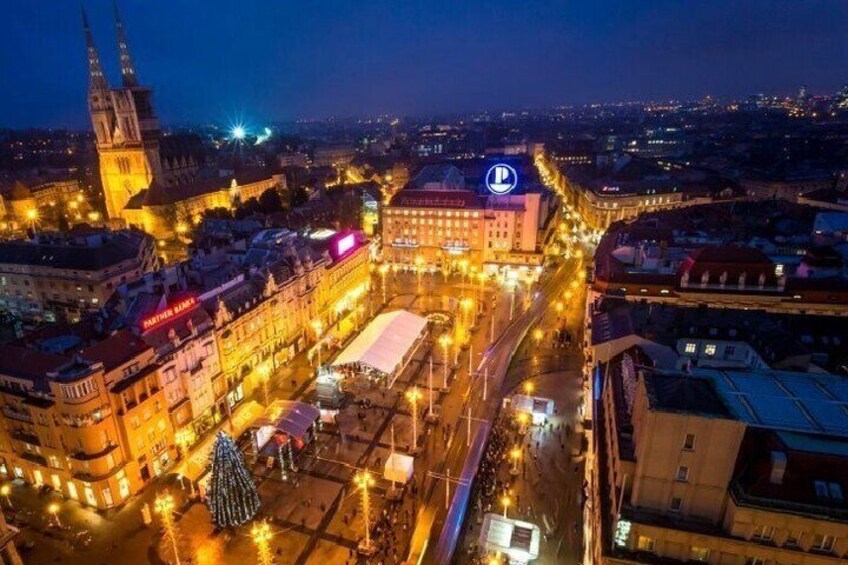 A cityscape at night with illuminated buildings, a prominent clock tower, and a Christmas tree in a square. 4