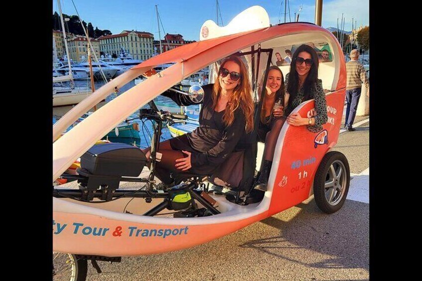 A small vehicle with three passengers, labeled "City Tour & Transport," parked near a marina with boats. 1