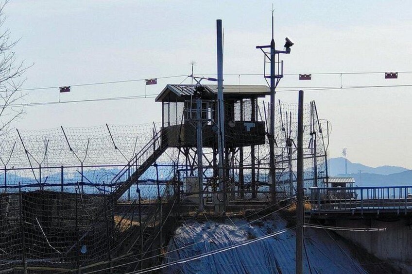 A watchtower with a metal staircase, surrounded by a wire fence, overlooking a body of water with mountains in the background. 3