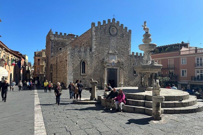 Un edificio de piedra histórico con una torre, una fuente y gente caminando en una plaza. 3
