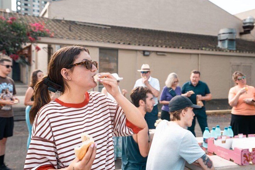 Una persona comiendo un sándwich en un evento al aire libre, con gente y mesas al fondo. 2