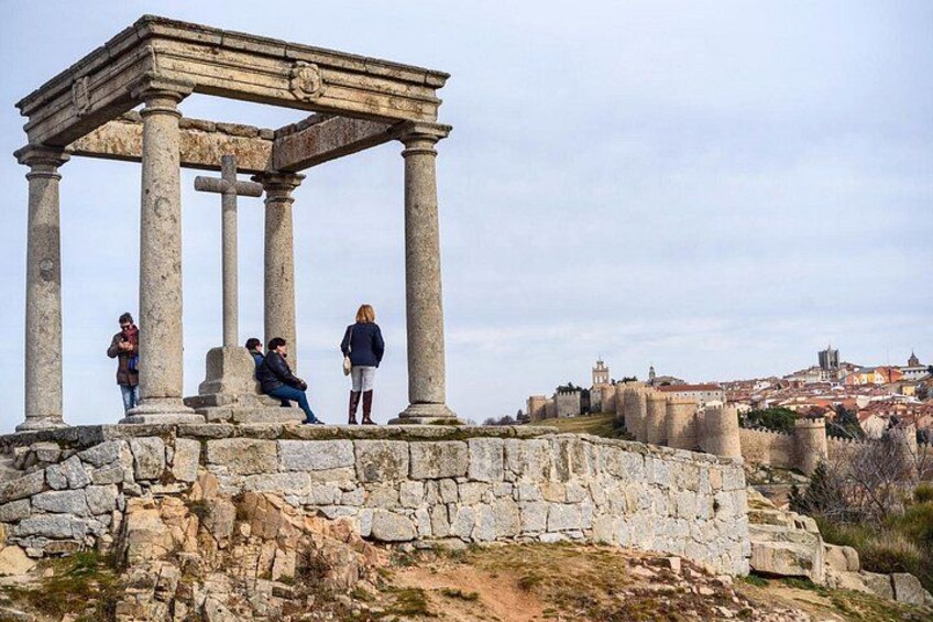A historical stone structure with columns, people standing around it, and a cityscape in the background. 5