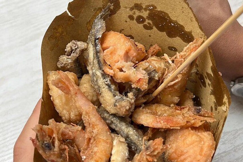 En hånd holder en æske med friturestegt fisk og skaldyr, herunder rejer og fisk, med spisepinde. 2