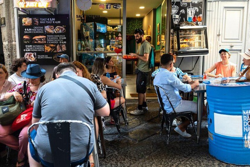 Des personnes sont assises à la terrasse d’un café, devant un panneau affichant le menu et un baril bleu. 2