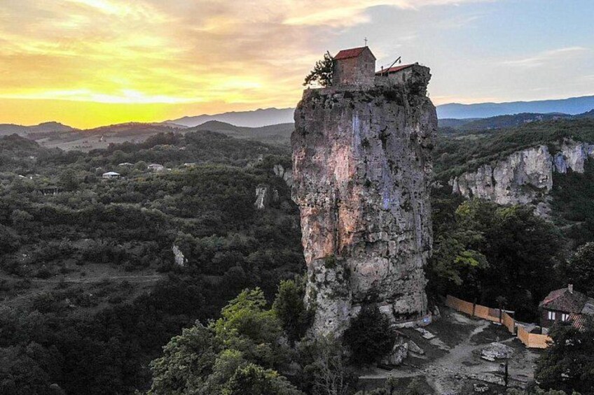 A tall rock formation with a small building on top, surrounded by dense forest and a clear sky. 4
