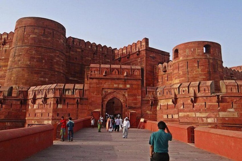 A large red fort with a central archway, a bridge leading to it, and people walking around. 4