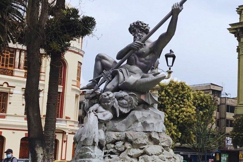 Eine Statue eines muskulösen Mannes, der einen Stab hält und auf einem Felsen sitzt, wobei ein Löwe zu seinen Füßen liegt, inmitten einer Stadt mit Gebäuden und Bäumen. 5